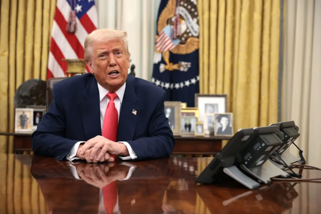 Donald Trump sentado no Salão Oval da Casa Branca durante pronunciamento oficial, com bandeiras e fotos familiares ao fundo.